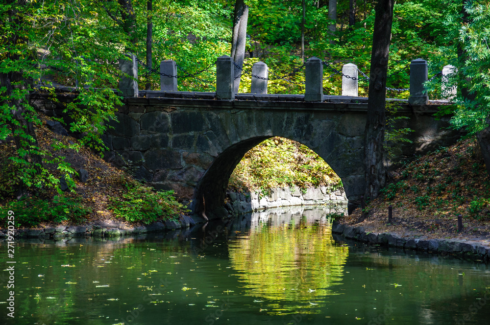 Ancient stone bridge with reflection in calm water surface of lake. Stone brick arch above pond. Landscape of Sofiyivka national park in Uman Ukraine. Autumn scene in Sofievka Uman park Ancient stone bridge with reflection in calm water surface of lake. Stone brick arch above pond. Landscape of Sofiyivka national park in Uman Ukraine. Autumn scene in Sofievka Uman park