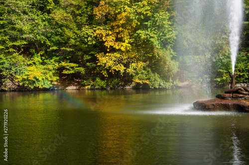 Water jet fountain on rock in center of lake with colorful fall trees reflected in calm water surface. Rainbow in water flow. Landscape of Sofiyivka park in Uman Ukraine. Autumn in Sofievka Uman park 