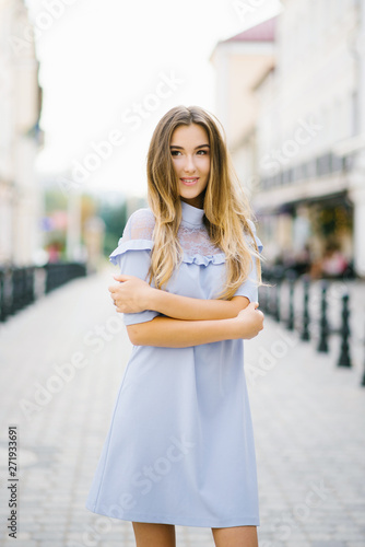 A beautiful young girl in a soft blue dress stands on the street of the city in the summer, smiling and happy. The concept of joy and happiness