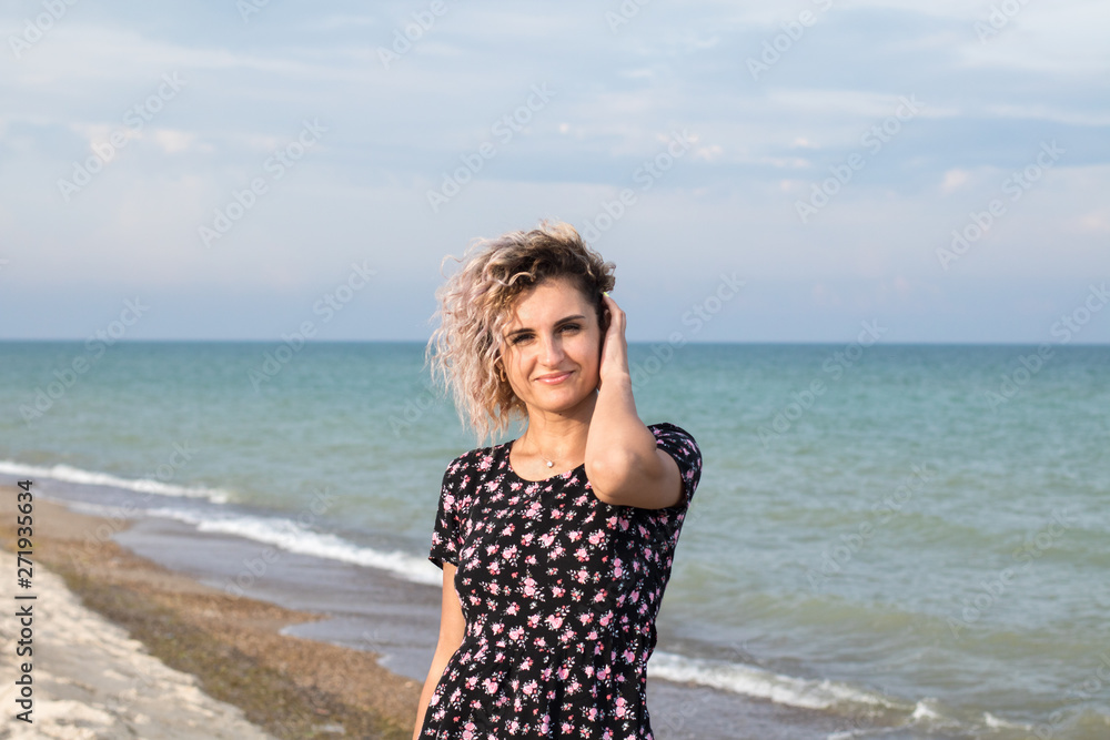 beautiful female with curly hairs in black dress in flowers is standing near water on the beach. Orange sunset light on the face beautiful female with curly hairs in black dress in flowers is standing near water on the beach. Orange sunset light on the face