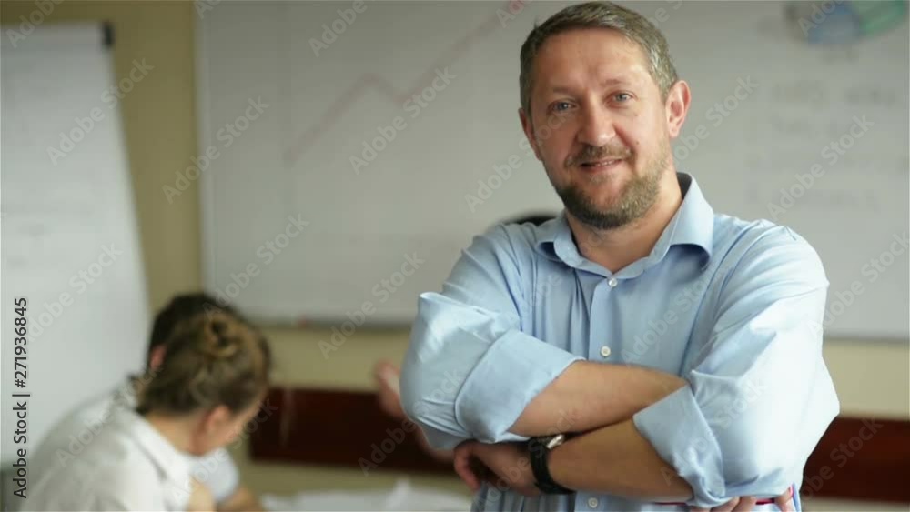 Happy businessman standing in the office with coworkers in the background working by the desk Happy businessman standing in the office with coworkers in the background working by the desk