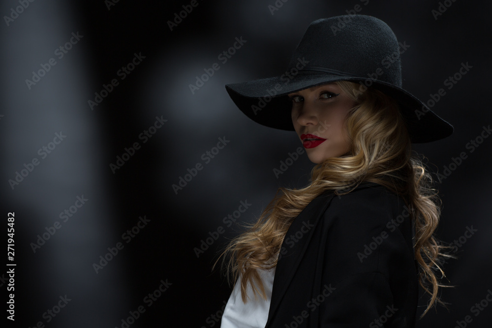 Side view of passionate girl with red lips and long curls looking at camera and posing in studio. Pretty model wearing black hat and smart suit standing on isolated background. Concept of beauty. Side view of passionate girl with red lips and long curls looking at camera and posing in studio. Pretty model wearing black hat and smart suit standing on isolated background. Concept of beauty.