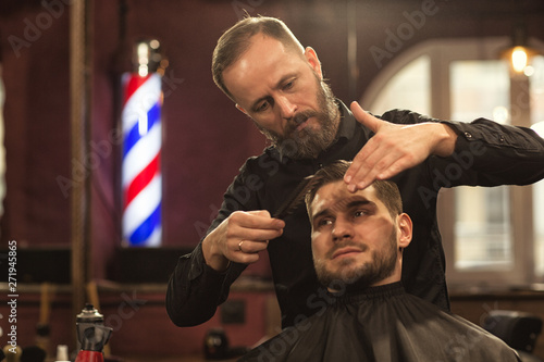 Front view of charming man in gown sitting in chair in barbersop during haircut. Serious bearded hairdresser in black shirt brushing forelock with comb and styling hair. Concept of male care.