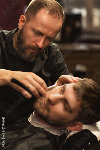 Selective focus of careful barber wearing black shirt in process of working in barbershop. Serious male master shaving beard of young client with razor. Concept of beard shape and care.