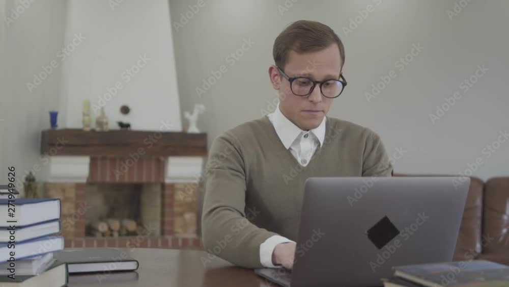 Young modestly dressed man in glasses sitting at the wooden table in the office, many books are on the table. Handsome nerd opening the laptop and start working