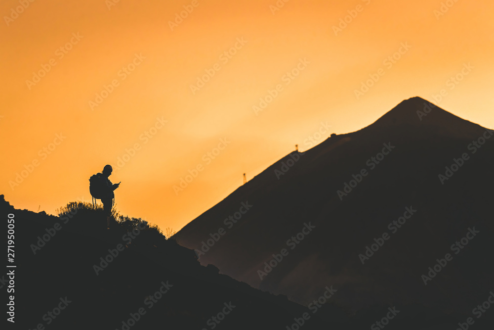 Silhouette of a young woman using smartphone after climbing.  Volcano Teide on background. Alone tourist with backpack enjoy nature and share with friends on vacation. Warm light of the golden hour