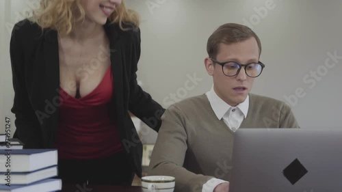 Portrait young adorable modestly dressed man in glasses sitting at table at home, working on laptop. Beautiful woman with deep neckline bends over and looking on screen, a man is confused. The nerd