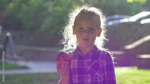 child bites apple in backlight, little girl dressed checkered shirt chews juicy ripe fruit and look on camera at nature
