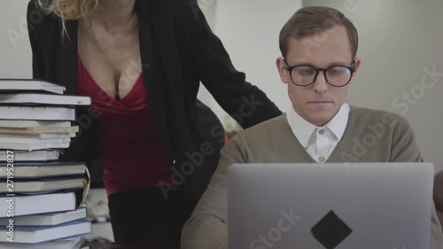 Young modestly dressed man in glasses sitting at table at home, working on laptop. Beautiful woman with deep neckline bends over and putting hand on his shoulder. The nerd trying to shake off the hand photo