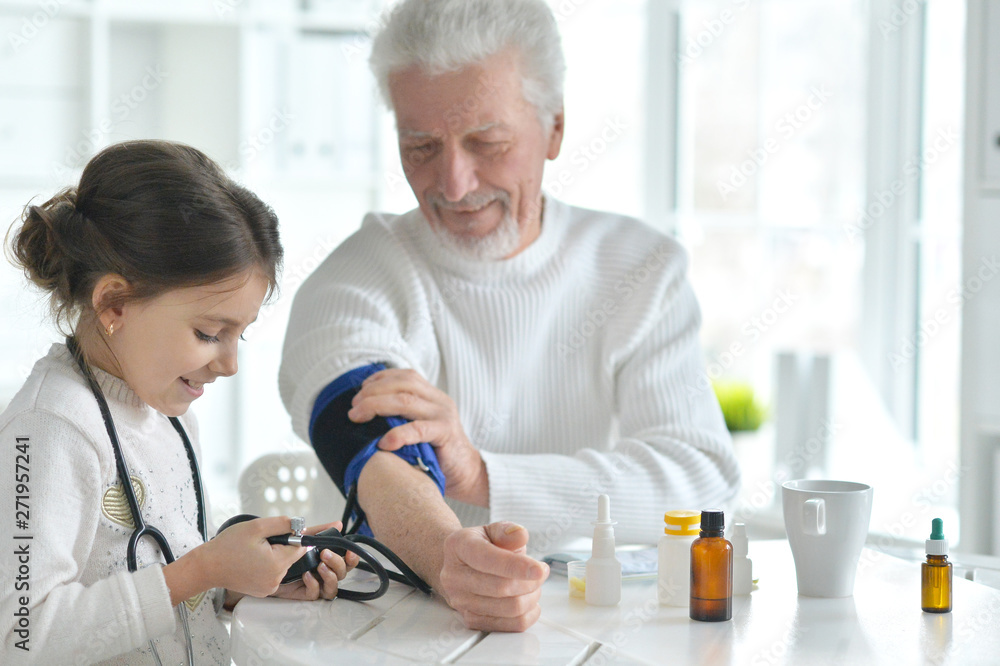 Fototapeta premium Portrait of granddaughter measuring blood pressure to her grandfather in room