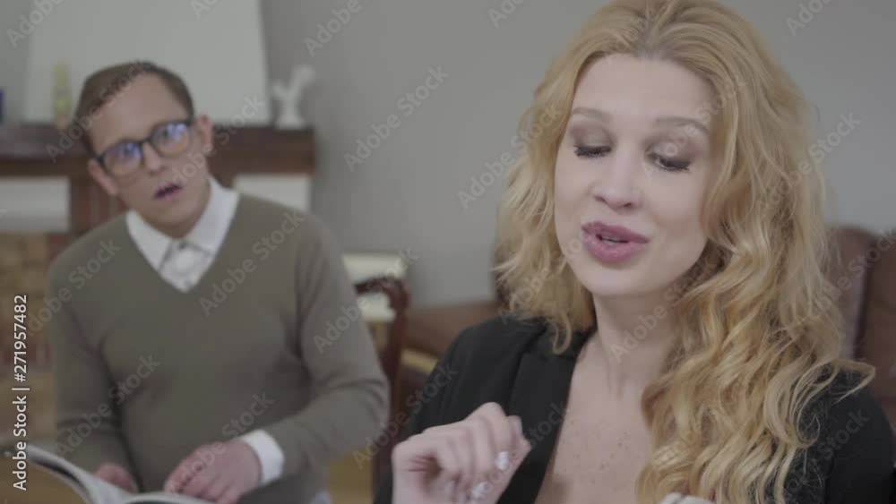 Portrait of beautiful blond woman reading aloud the book in the foreground while modestly dressed man looking at the lady on the background. The nerd and hot woman working together Portrait of beautiful blond woman reading aloud the book in the foreground while modestly dressed man looking at the lady on the background. The nerd and hot woman working together