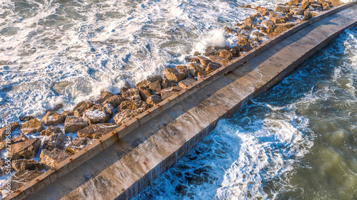 Aerial view on a stone pier against which the force of the sea is raging with high and strong waves. On the construction in the middle of the water there is nobody.Waves banging create the white foam.
