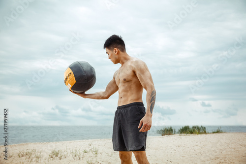 Young healthy man athlete doing exercise with ball at the beach. Signle male model shirtless training air at the river side in sunny day. Concept of healthy lifestyle, sport, fitness, bodybuilding. photo