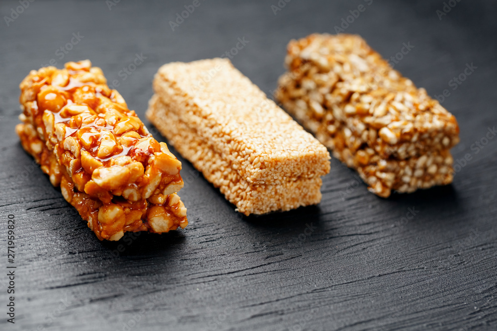 Cereal granola bar with peanuts, sesame and sunflower seeds on a cutting board on a dark stone table. View from above. Three Assorted Bars Cereal granola bar with peanuts, sesame and sunflower seeds on a cutting board on a dark stone table. View from above. Three Assorted Bars