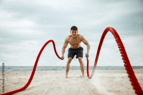 Young healthy man athlete doing exercise with the rope at the beach. Signle male model shirtless training air at the river side in sunny day. Concept of healthy lifestyle, sport, fitness, bodybuilding photo