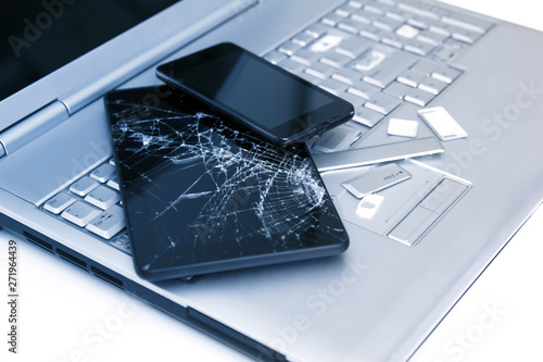 A silver laptop with a broken keyboard, tablet with a cracked display and black phone. A close-up picture of part of broken laptop and cracked screen on a tablet.