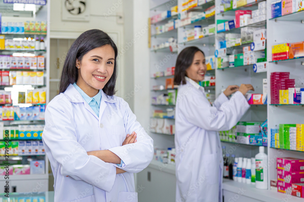 Smiling asian pharmacists crossing arms in front of the modern drugstore with her colleague. Health care and medical concept.