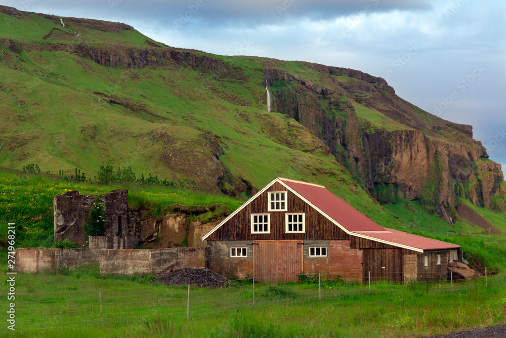 The wooden house with red roof back of the fences under the green rocky cliffs surrounded with green grasses The wooden house with red roof back of the fences under the green rocky cliffs surrounded with green grasses