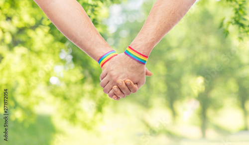 lgbt, same-sex relationships and homosexual concept - close up of male couple wearing gay pride rainbow awareness wristbands holding hands over green natural background © Syda Productions