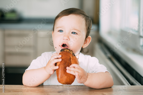 a little girl in the kitchen during the day eating a bun with poppy seeds is very appetizing