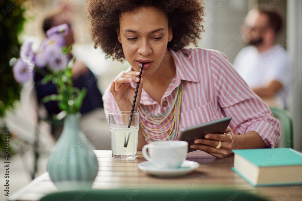 Gorgeous mixed race woman in pink striped dress drinking lemonade and using tablet while sitting in cafe.