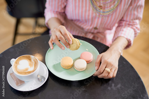 Top view of mixed race woman in pink striped dress holding cookie while sitting in pastry shop. On table are coffee and plate with cookies.