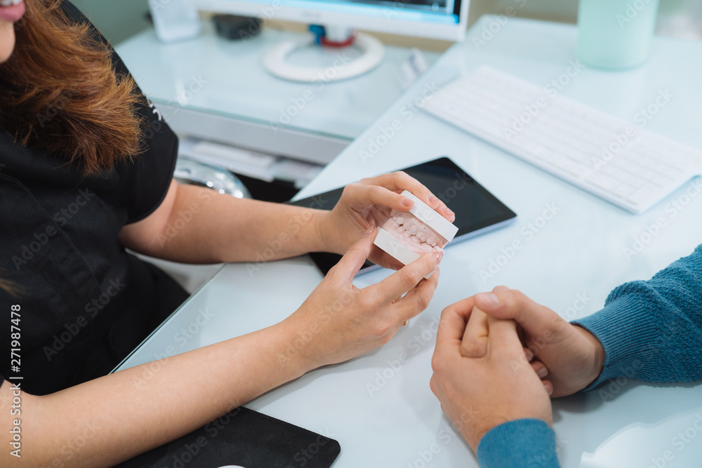 Female dentist holding tooth mold, explaining something to her patient during medical consultation at her clinic. Orthodontist showing her client denture. Experience, medical treatment concept. Female dentist holding tooth mold, explaining something to her patient during medical consultation at her clinic. Orthodontist showing her client denture. Experience, medical treatment concept.