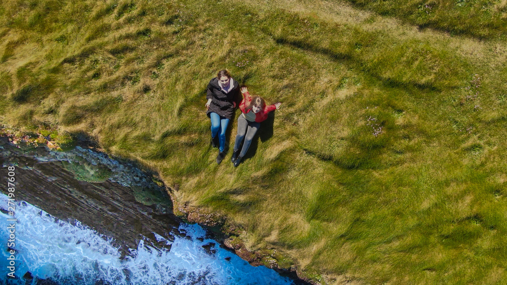 Two girls lying in the grass at the cliffs of the Irish coast - drone view from above - travel photography