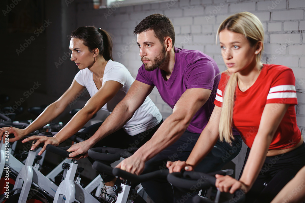 Group of smiling friends at gym exercising on stationary bike. Happy cheerful athletes training on exercise bike. Young men and woman working out at a class in the gym.