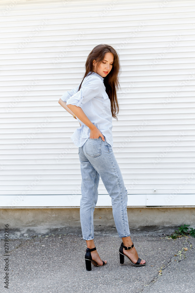 Young stylish woman wearing blue shirt, blue cropped denim jeans and black  high heel sandals posing against white street wall. Trendy casual outfit.  Street fashion. Stock Photo | Adobe Stock