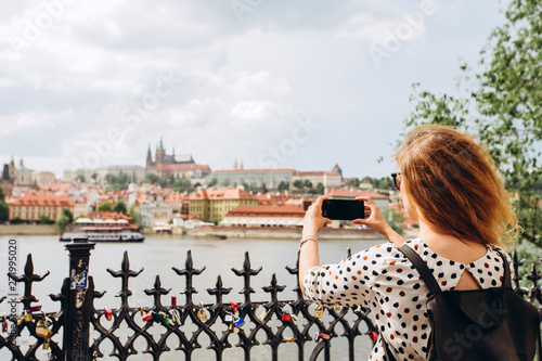 A girl with a backpack makes a photo on the phone of the Charles Bridge of Prague. Tourist girl takes pictures of Prague Castle. A woman holds a smartphone in her hands.