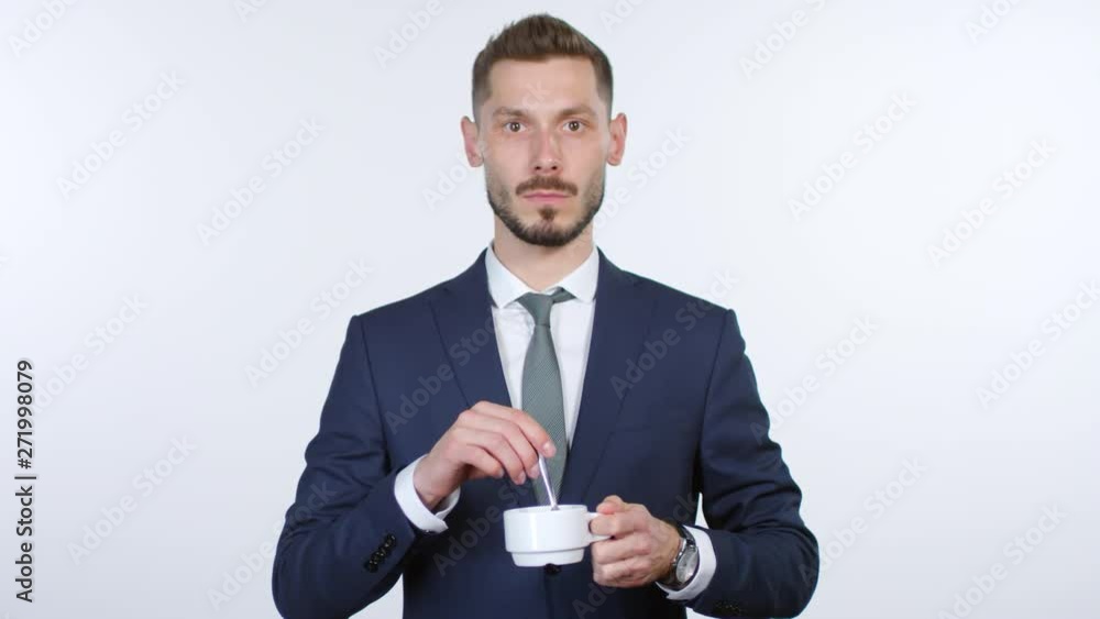 Portrait of emotionless businessman in suit standing isolated on grey background and stirring coffee in cup while staring at camera Portrait of emotionless businessman in suit standing isolated on grey background and stirring coffee in cup while staring at camera