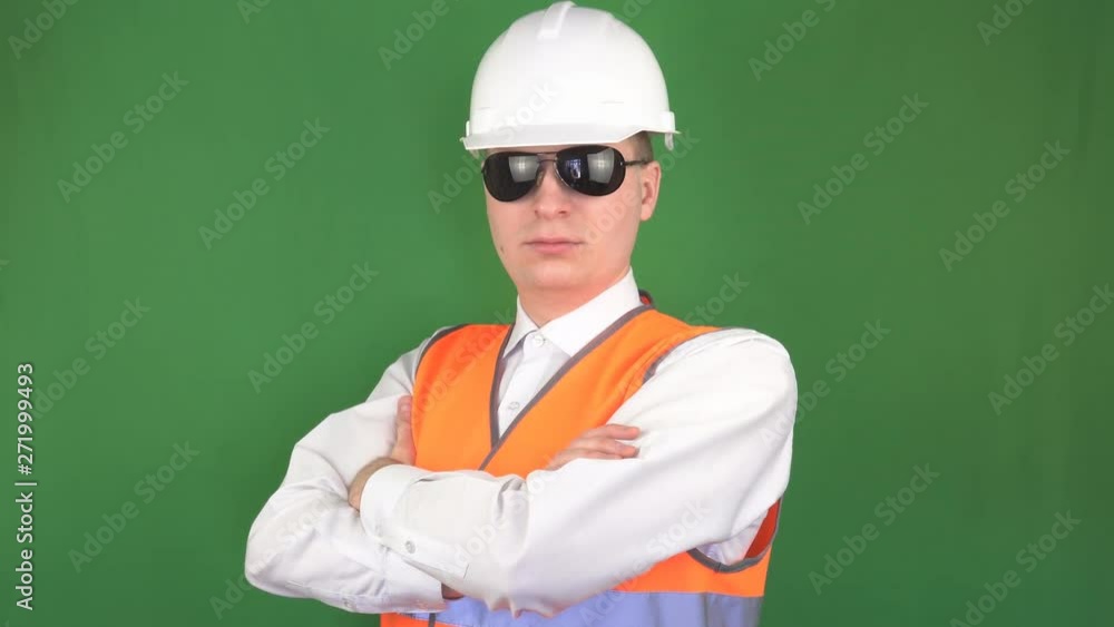 A young caucasian man in black glasses with a protective white helmet and a signal gilet looking into the camera, green background, hromakey, prevention of accidents A young caucasian man in black glasses with a protective white helmet and a signal gilet looking into the camera, green background, hromakey, prevention of accidents
