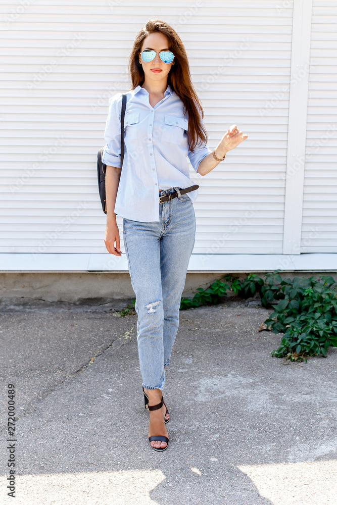 Young stylish woman wearing blue shirt, blue cropped denim jeans, black  high heel sandals, black backpack and sunglasses posing against white  street wall. Trendy casual outfit. Street fashion. Stock Photo | Adobe