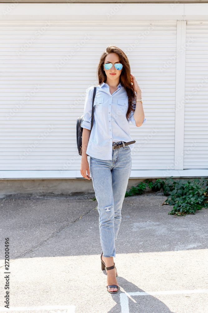 Young stylish woman wearing blue shirt, blue cropped denim jeans, black  high heel sandals, black backpack and sunglasses posing against white  street wall. Trendy casual outfit. Street fashion. foto de Stock