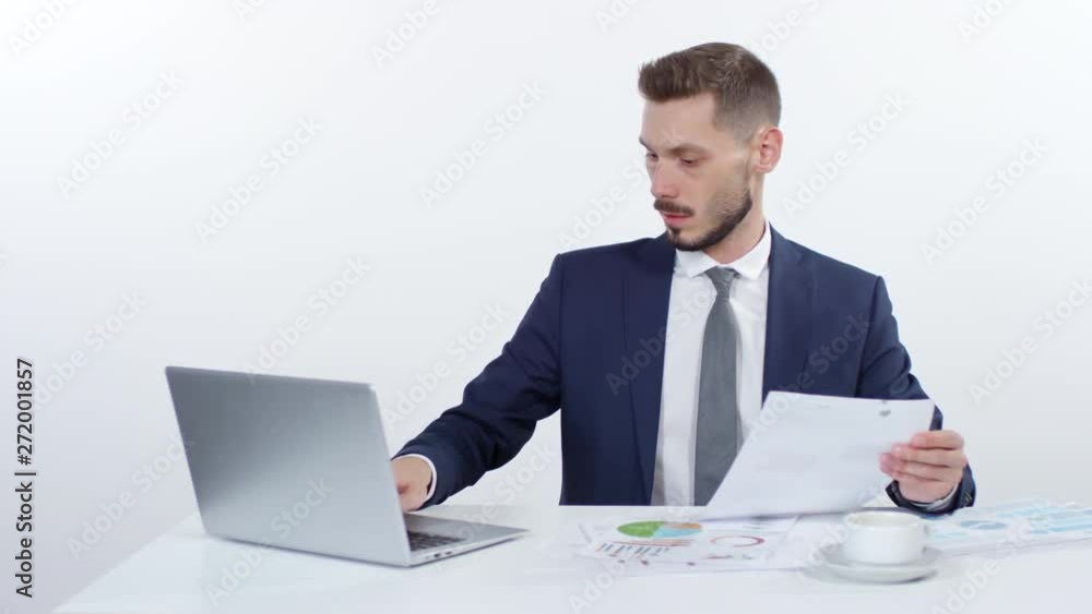 Medium shot of businessman in suit sitting at desk and typing on laptop while going over documents, then drinking coffee and smiling Medium shot of businessman in suit sitting at desk and typing on laptop while going over documents, then drinking coffee and smiling