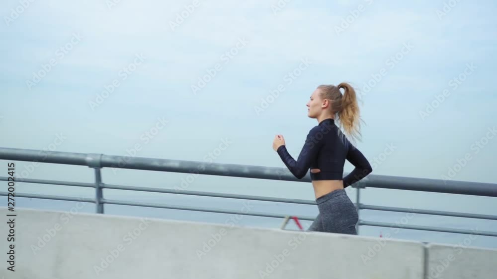 Tracking left of determined young woman jogging on bridge road over water during marathon training, side view slow motion shot Tracking left of determined young woman jogging on bridge road over water during marathon training, side view slow motion shot