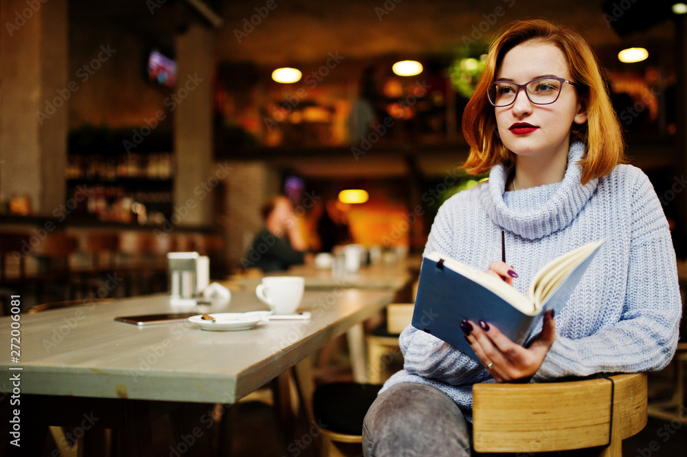 Cheerful young beautiful redhaired woman in glasses sitting at her working place on cafe reading something at her notebook. Cheerful young beautiful redhaired woman in glasses sitting at her working place on cafe reading something at her notebook.
