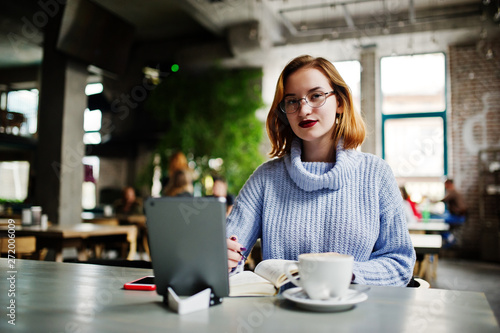 Cheerful young beautiful redhaired woman in glasses using her phone, touchpad and notebook while sitting at her working place on cafe with cup of coffee.