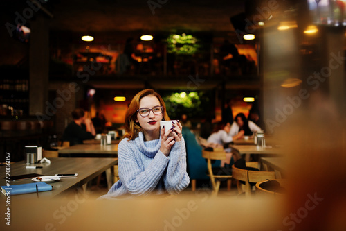 Cheerful young beautiful redhaired woman in glasses sitting at her working place on cafe and drinking coffee.