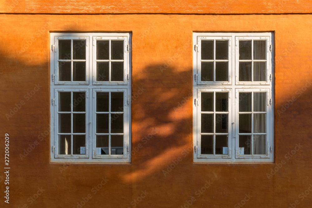 Two old style white wooden windows on a bright orange wall, partly covered by a shadow.