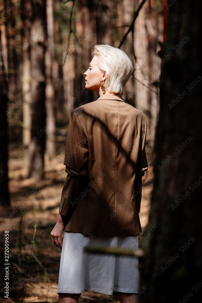 Fototapeta premium pensive blonde woman in brown jacket standing in forest