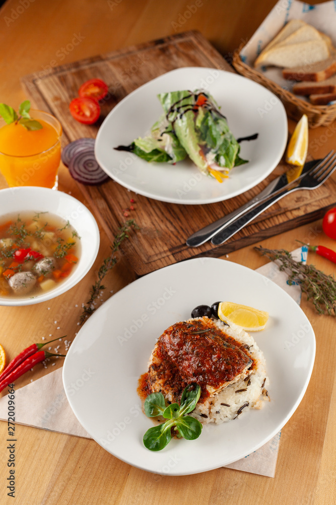 Lunching out in a restaurant: fish with steamed rice, light vegetable soup with meatballs and vegetarian spring roll on a dark background