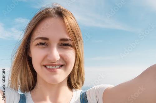 Young smiling girl dressed in a white T-shirt, a selfie on the background of the sea on a warm summer day
