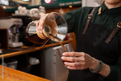 Female barista is pouring black coffee, made in chemex, into a glass, standing in front of coffee ouse interior photo