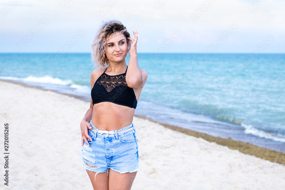 beautiful young woman with curly hairs in denim shorts and swimsuit is happy to be on the beach near the ocean. Ocean and grass on the background beautiful young woman with curly hairs in denim shorts and swimsuit is happy to be on the beach near the ocean. Ocean and grass on the background