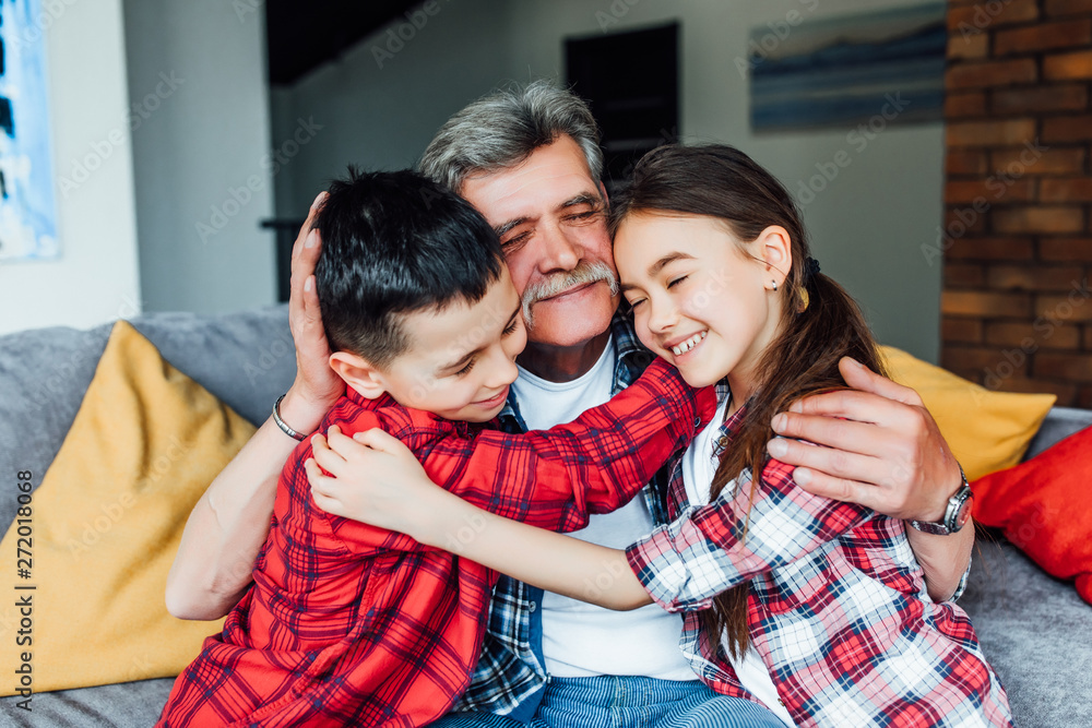 Relax. Nice cheerful grandfather smiling and sitting on the couch with his grandchildren and hugging their. Relax. Nice cheerful grandfather smiling and sitting on the couch with his grandchildren and hugging their.