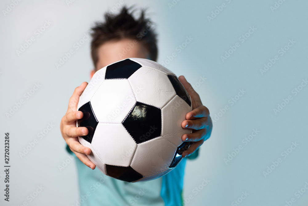 boy in a blue t-shirt holding a soccer ball in his hands obscuring his head on a blue background