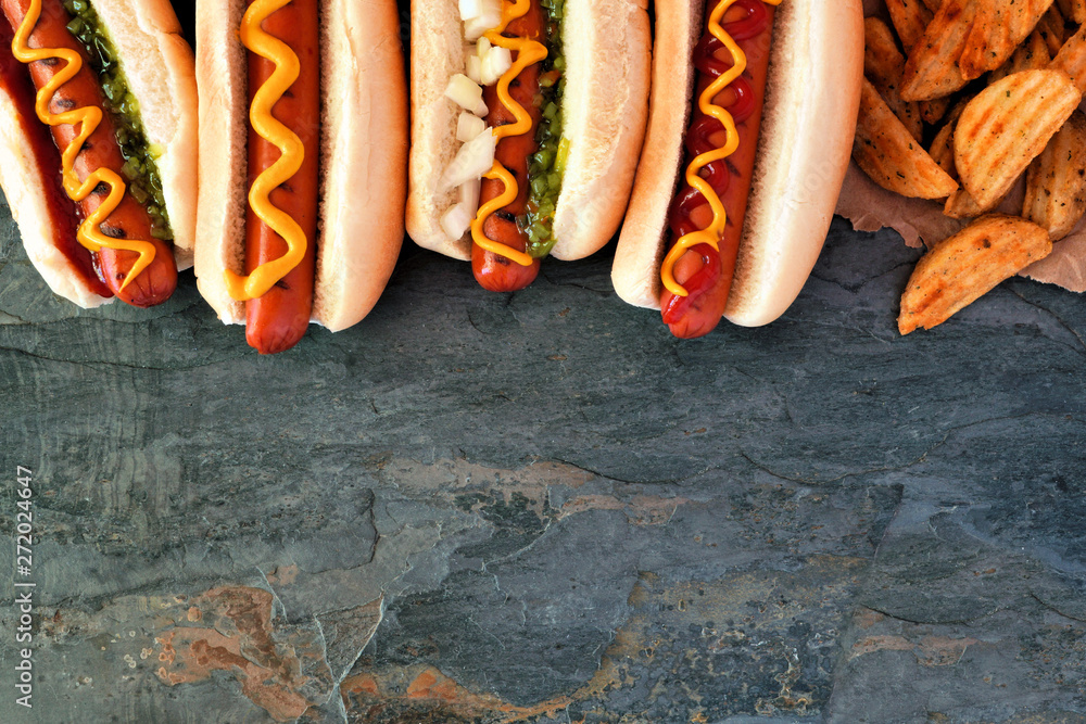 Hot dogs with toppings and potato wedges. Top border, overhead view on a dark stone background with copy space. - obrazy, fototapety, plakaty 