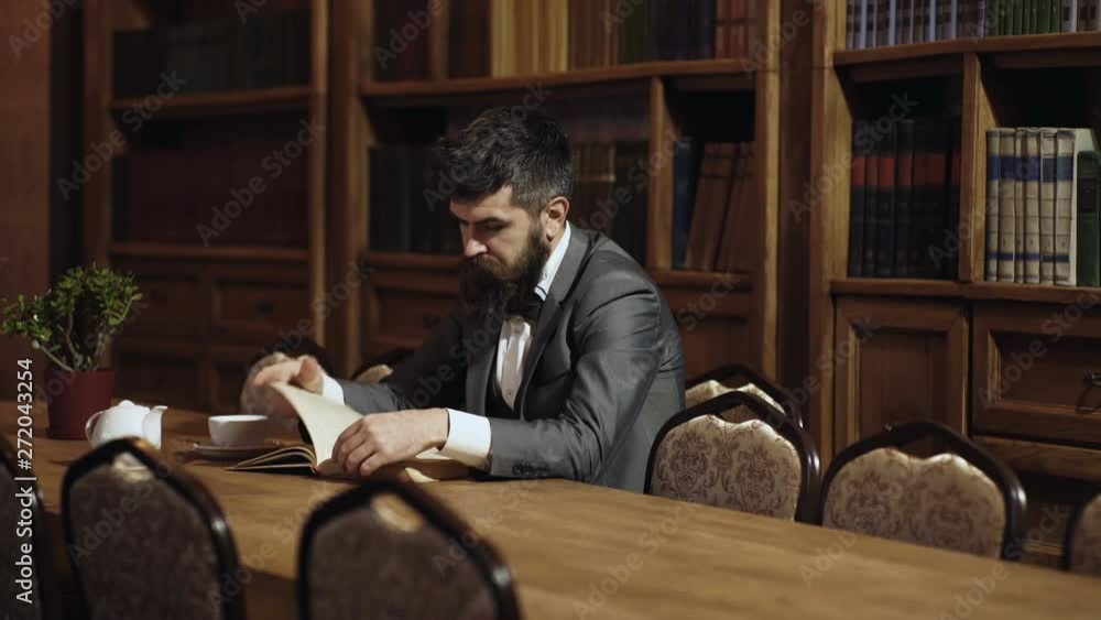 Man reading a book during coffee break. Man in classic suit sits in vintage interior, library, book shelves on background. Aristocratic lifestyle concept. Aristocrat on thoughtful face reading book. Man reading a book during coffee break. Man in classic suit sits in vintage interior, library, book shelves on background. Aristocratic lifestyle concept. Aristocrat on thoughtful face reading book.